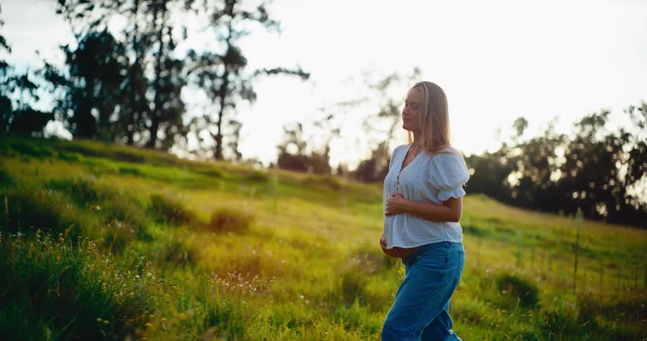 A beautiful pregnant woman walks through a grassy field in the golden light of sunset, smiling and peacefully connecting with her baby