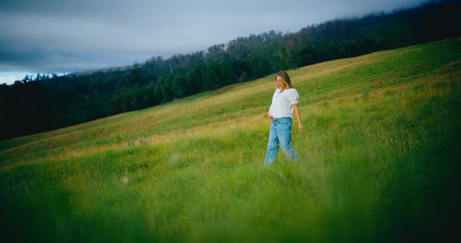 A happy pregnant woman gently cradles her belly while walking through a lush green hillside meadow at sunset