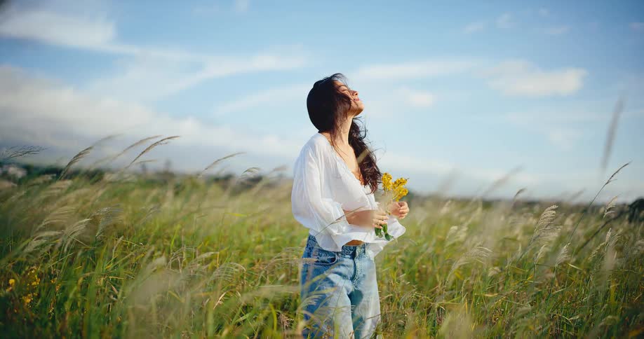 A beautiful young woman with a bouquet of yellow wildflowers enjoys the sun and breeze in a grassy field