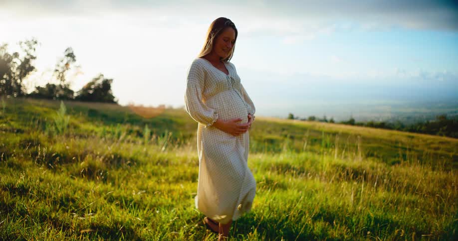 A serene pregnant woman in a white dress walks through a golden, sunlit grassy field while holding her belly