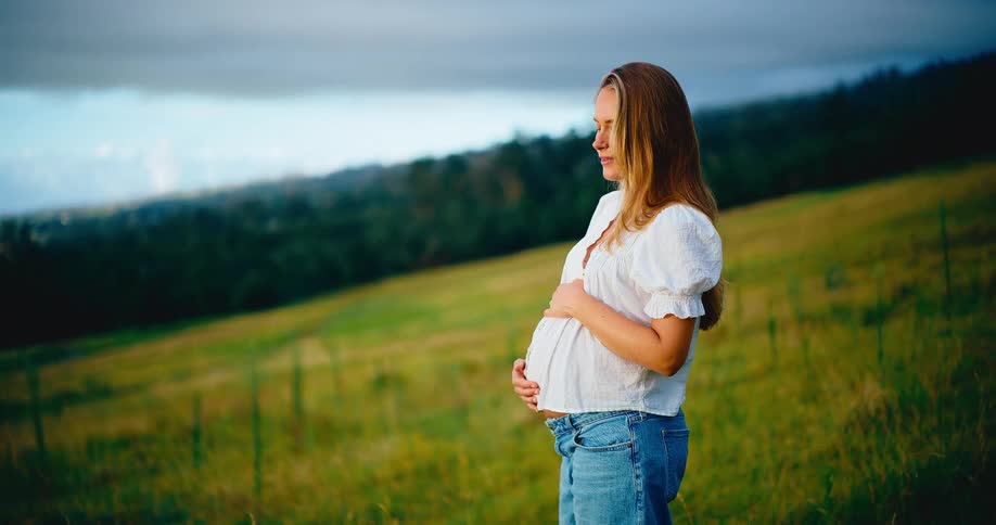 A thoughtful pregnant woman gently holds her belly while standing in a beautiful green field, enjoying a moment of peace and reflection