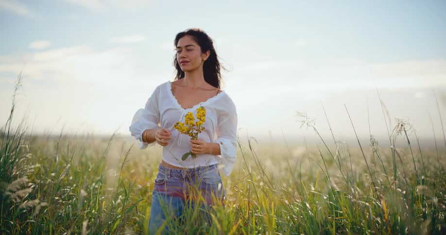A beautiful young woman holding yellow wildflowers stands peacefully in a sunlit field of tall grass as a gentle breeze blows