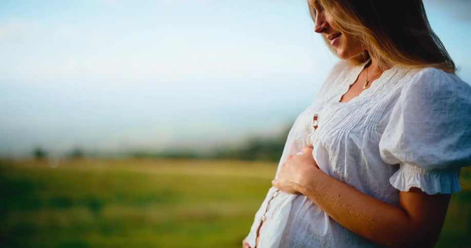 A beautiful pregnant woman smiles as she gently holds her baby bump while standing in a sunlit green field