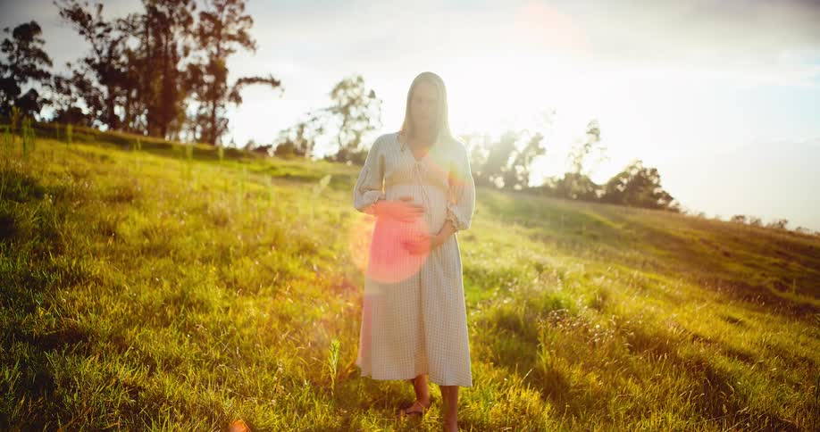 A beautiful pregnant woman stands in a golden field at sunset, lovingly holding her belly and smiling