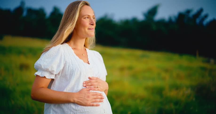 A beautiful pregnant woman in a white dress smiles serenely while holding her belly in a golden field at sunset