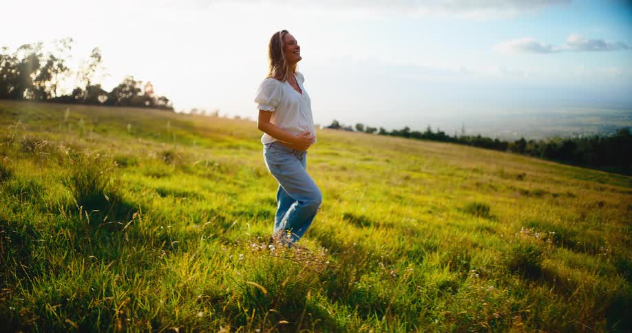 A smiling pregnant woman walks through a beautiful green field during a warm sunset, caressing her baby bump