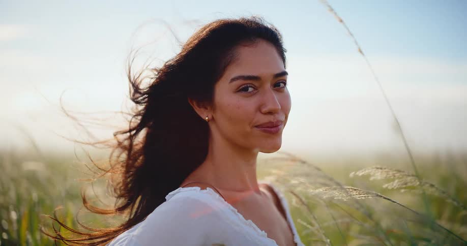 A serene woman with her hair blowing in the wind smiles and closes her eyes in a sunlit field of tall grass