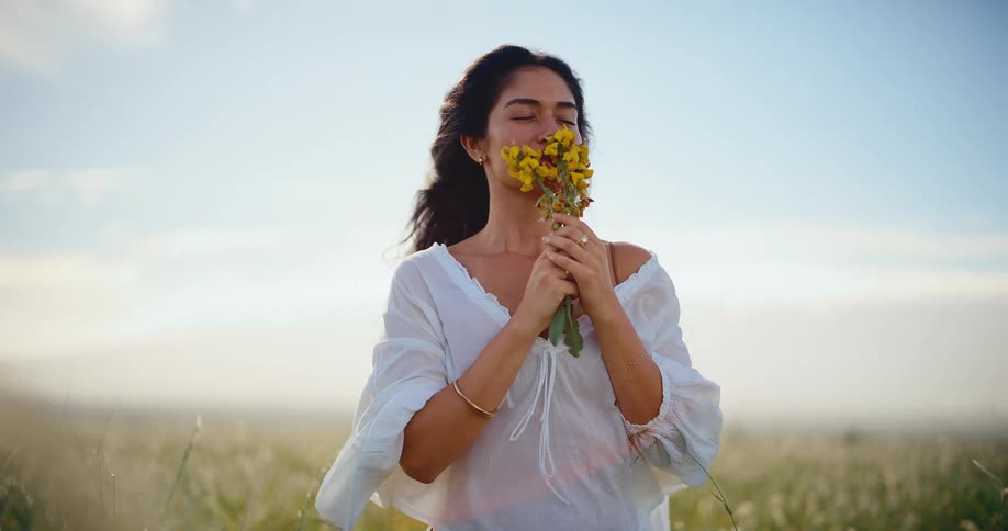 A beautiful woman smells a bouquet of yellow wildflowers while standing in a breezy, sunlit field in slow motion