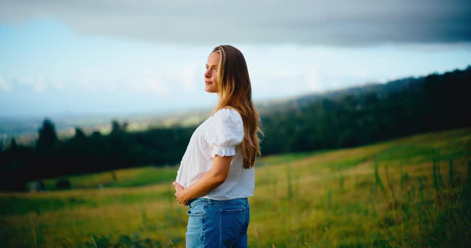 A content pregnant woman holds her baby bump while standing peacefully in a lush green field enjoying the view