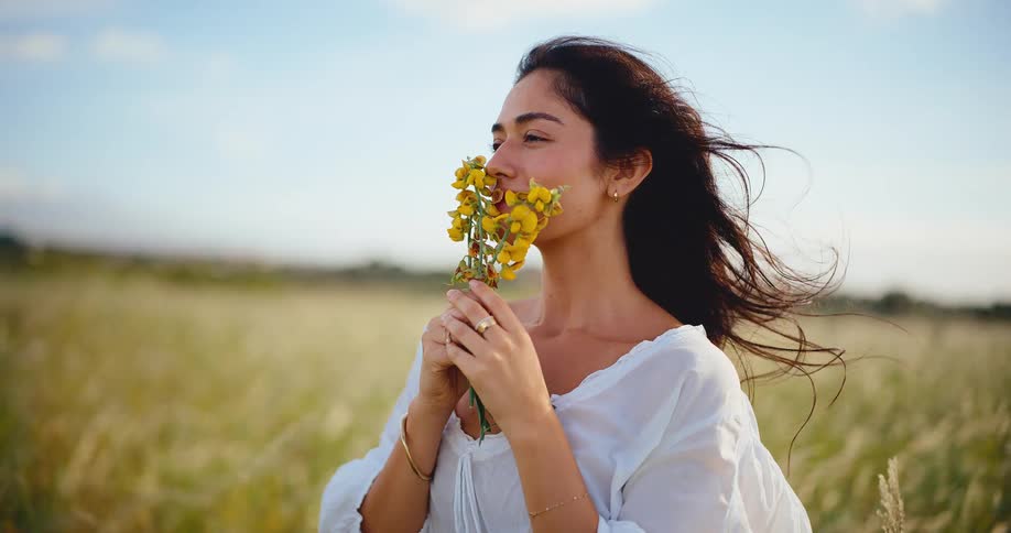 Slow motion of a serene young woman in a white blouse smelling a bouquet of yellow wildflowers while standing in a breezy, sunlit field
