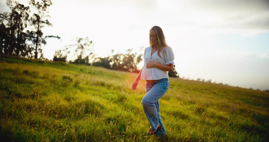 A happy pregnant woman gently walks through a golden sunlit field, tenderly holding her baby bump