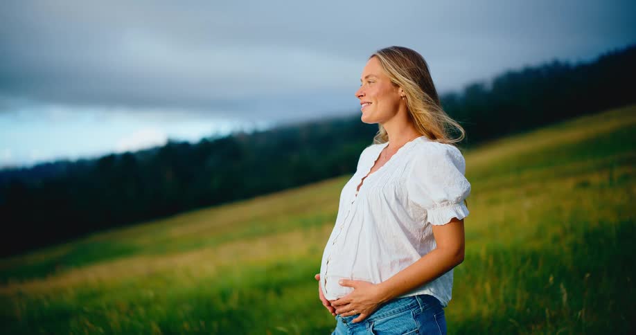 A serene and beautiful pregnant woman stands in a green field at sunset, smiling as she holds her baby bump