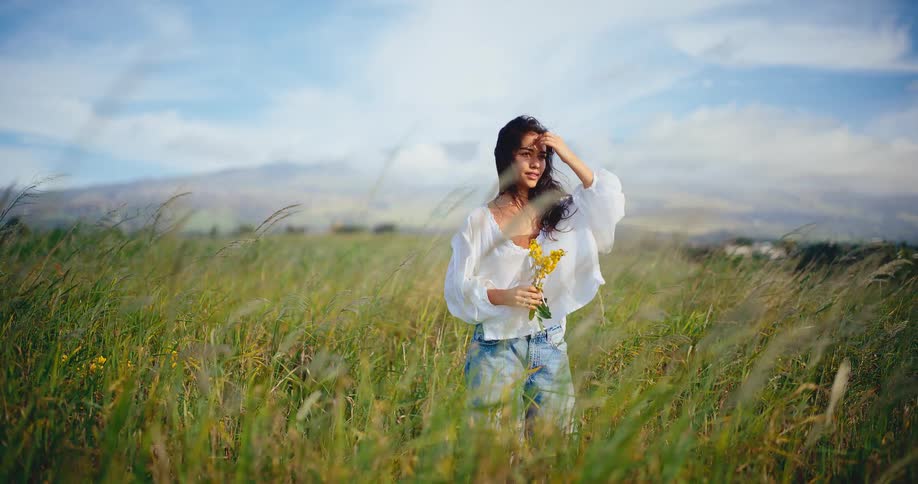A serene young woman holds yellow wildflowers while standing in a breezy green field, lifting her face to the sun