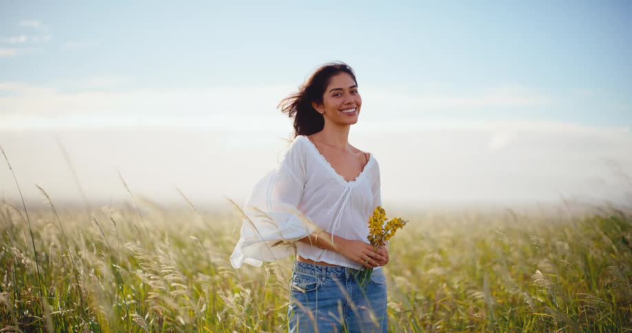 Portrait of a beautiful woman holding yellow wildflowers smiling in a windy field at sunset