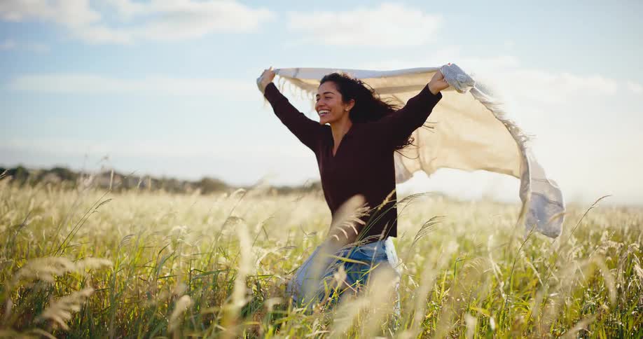 A joyful young woman runs through a sunny, grassy field in slow motion, holding a blanket that billows in the wind