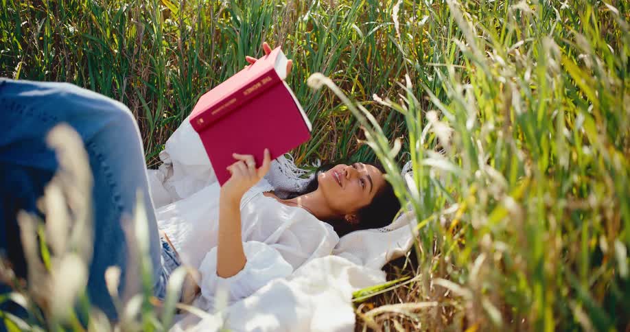 A happy young woman relaxes on a blanket in a field of tall, sunlit grass while reading a book