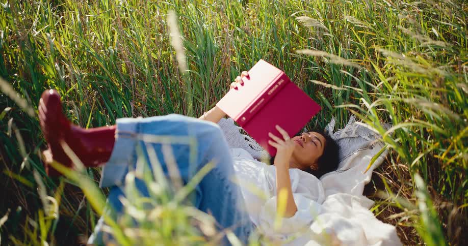 A young woman relaxes in a sunny field of tall grass, reading a book as she lies on a blanket