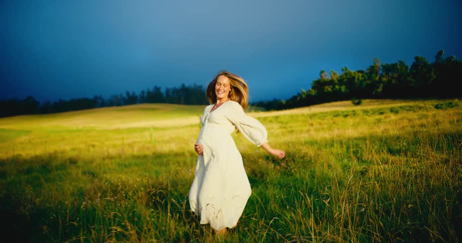 Serene and happy pregnant woman walks through a sunlit grassy meadow in slow motion, celebrating new life under a dramatic sky