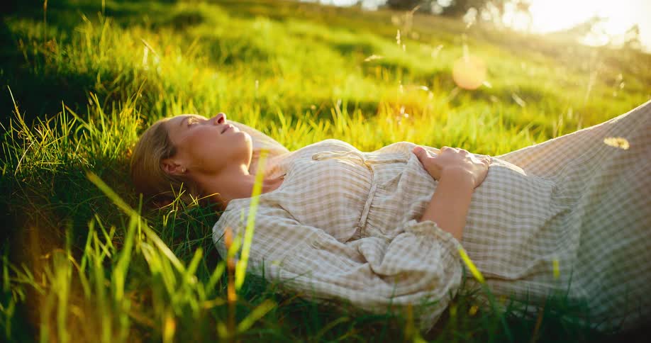 A serene pregnant woman lies in a lush green meadow, peacefully basking in the warm glow of the sun