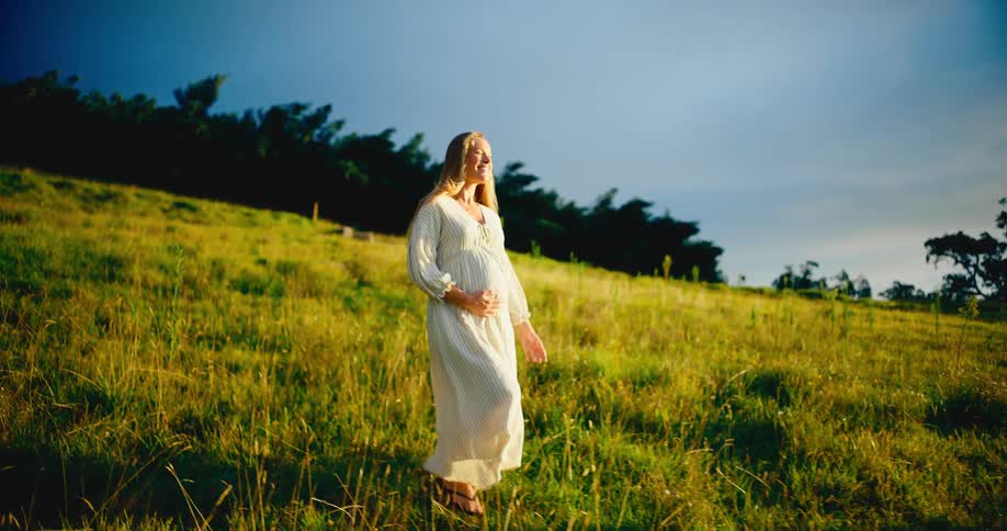 A serene pregnant woman in a flowing white dress smiles as she walks through a sunlit grassy field in slow motion