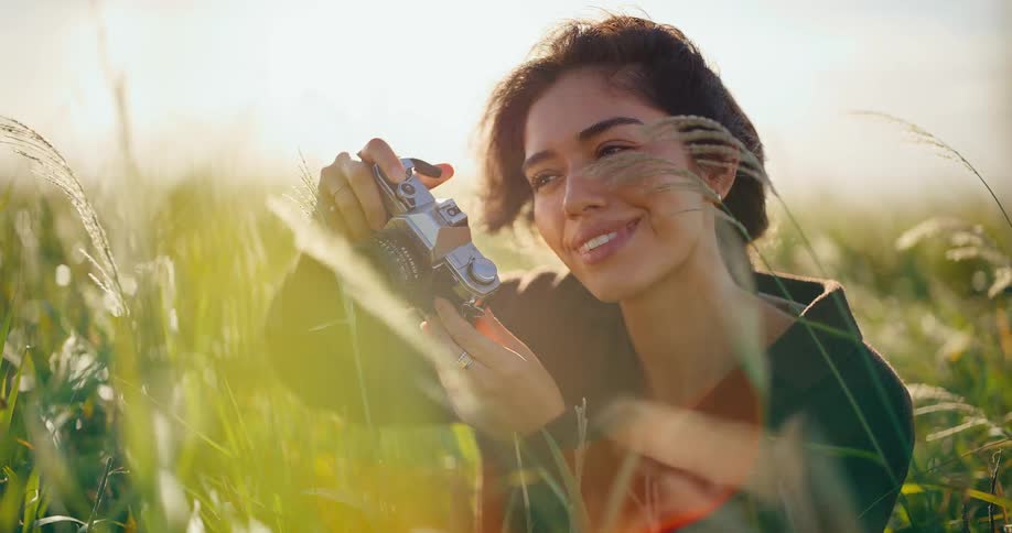 A smiling young woman uses a vintage film camera to take pictures in a beautiful sunlit field of tall grass