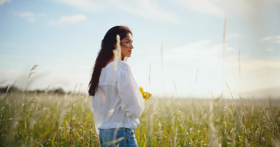 A serene young woman with long dark hair smells yellow wildflowers in a sunlit field of tall grass in slow motion