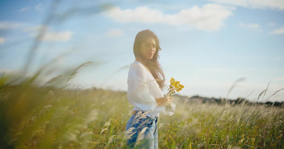 Slow motion portrait of a beautiful woman with a bouquet of yellow wildflowers enjoying the breeze in a sunlit meadow