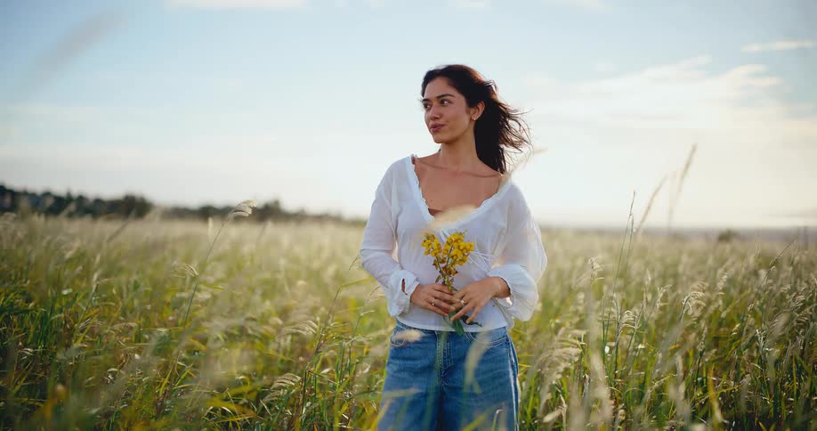 Beautiful young woman holding yellow flowers stands peacefully in a golden field at sunset