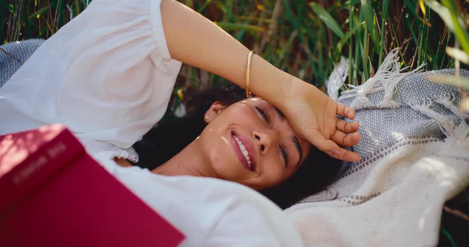 A happy young woman with a book lies peacefully on a blanket in a sunny meadow, with dappled sunlight on her face