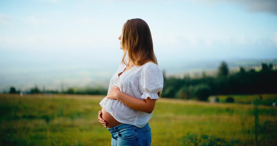 An expectant mother gently cradles her baby bump while enjoying a moment of serenity in a lush green meadow at golden hour