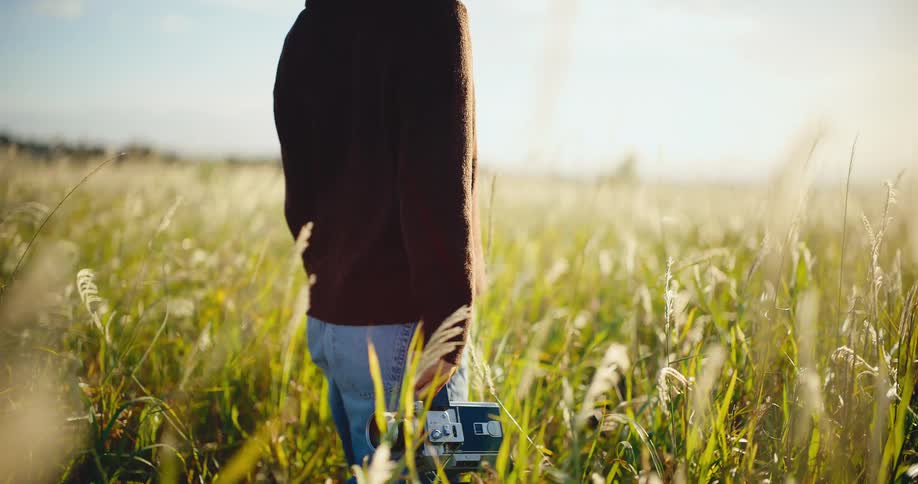 A filmmaker walks through a sun-drenched field of tall grass with a vintage camera during golden hour