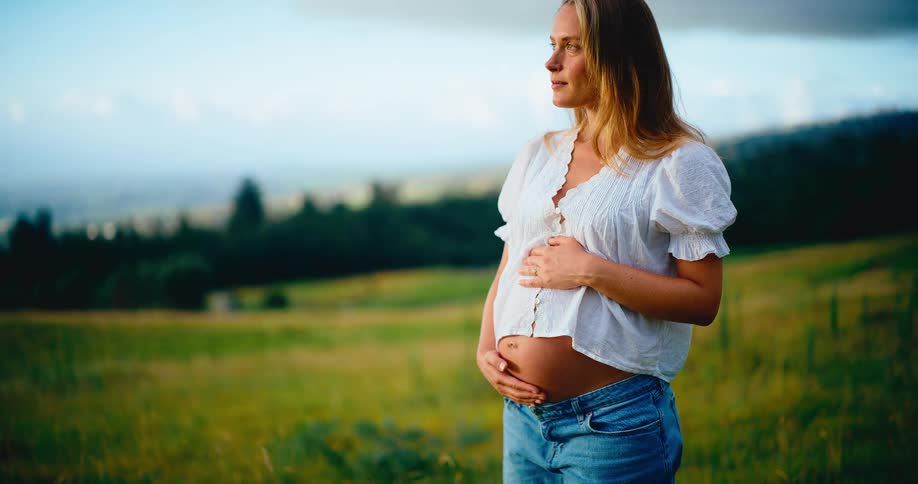 A serene pregnant woman embraces her belly while standing in a peaceful, sunlit green field