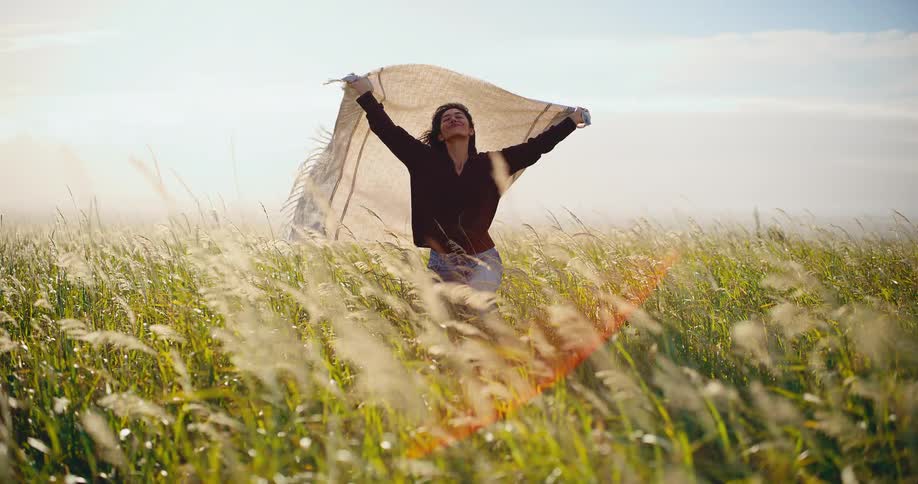 Slow motion shot of a happy woman standing in a tall grassy field, holding a blanket up in the wind with her arms outstretched