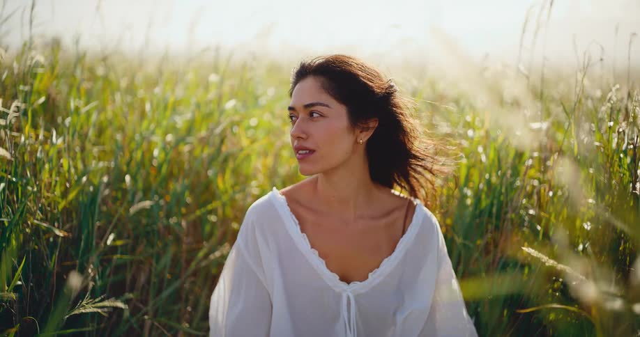 A serene young woman enjoys the golden hour sunlight while sitting peacefully in a field of tall green grass