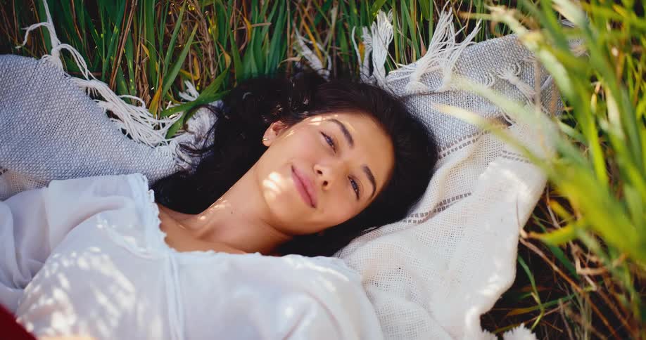 A smiling woman lowers her book to reveal her face as she lies peacefully on a blanket in a sunlit grassy meadow