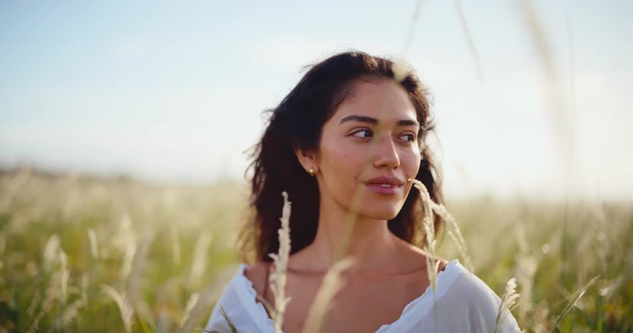 A beautiful brunette woman with her hair blowing in the wind smiles serenely while standing in a sunlit field of tall grass