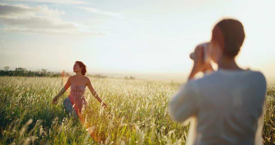 A woman uses a vintage camera to film her friend posing in a sun-drenched grassy field during golden hour