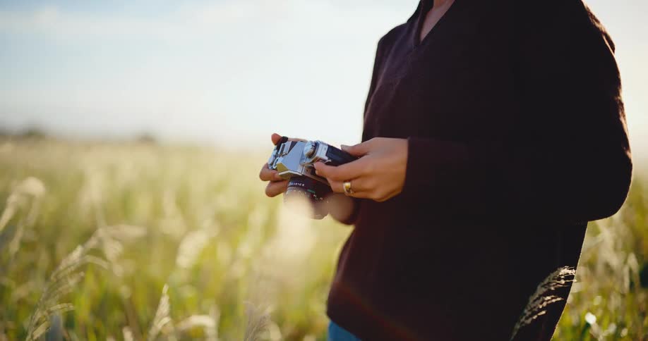 A woman with a vintage film camera adjusts its settings while standing in a sun-drenched field of tall grass