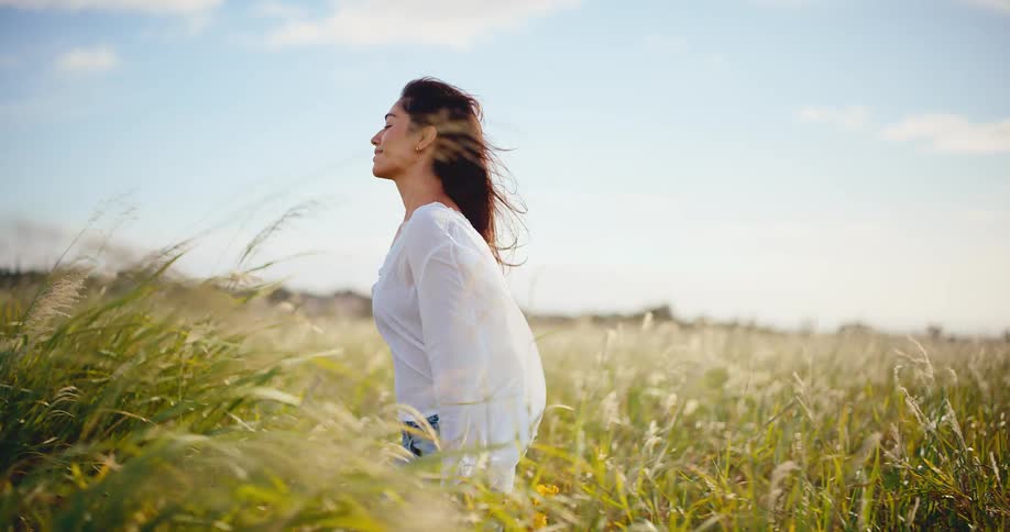 A beautiful young woman in a white blouse smiles peacefully as she walks through a sunlit field of tall grass with the wind in her hair