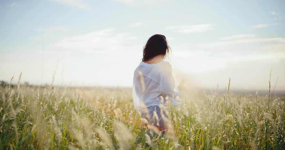 A beautiful woman smiles while holding wildflowers and standing in a golden sunlit field as the wind blows her hair