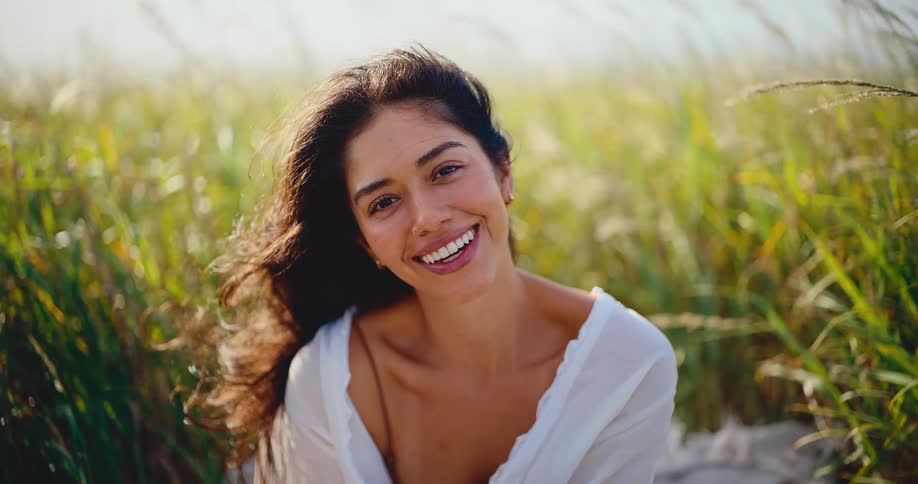 A beautiful young woman reading a book in a sunlit field of grass smiles radiantly at the camera