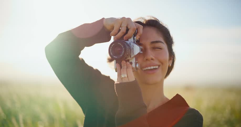 A joyful photographer smiles while taking a picture with her vintage film camera in a sun-drenched field in slow motion