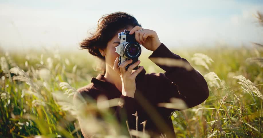 A beautiful young woman with a vintage film camera takes a picture and smiles serenely in a sunlit field of tall grass
