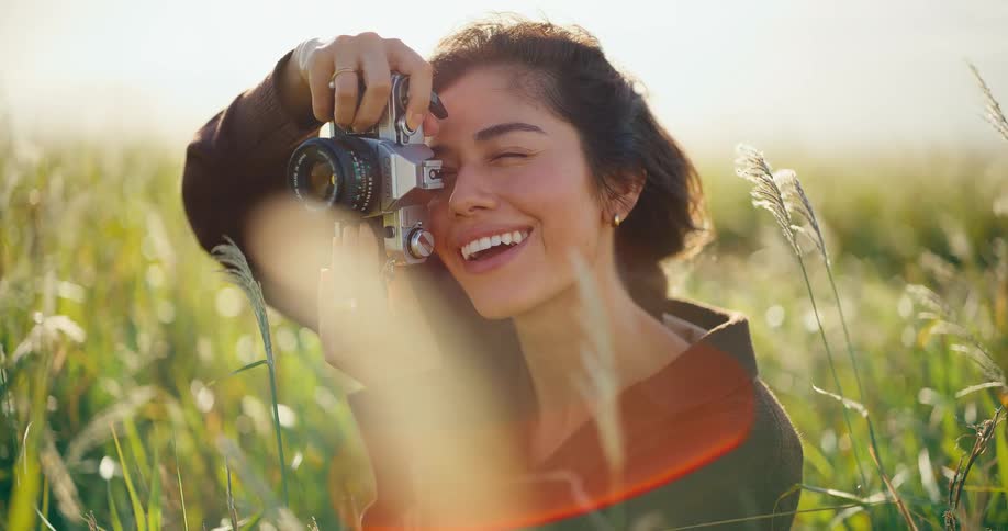 A joyful young female photographer smiles while taking pictures with a classic film camera in a sun-drenched meadow