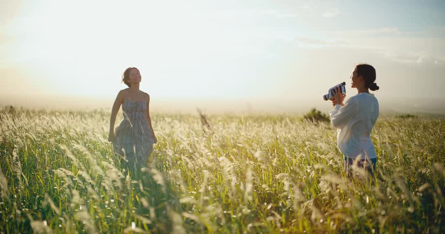 A woman films her friend with a vintage 8mm camera in a golden sunlit field of tall grass blowing in the wind