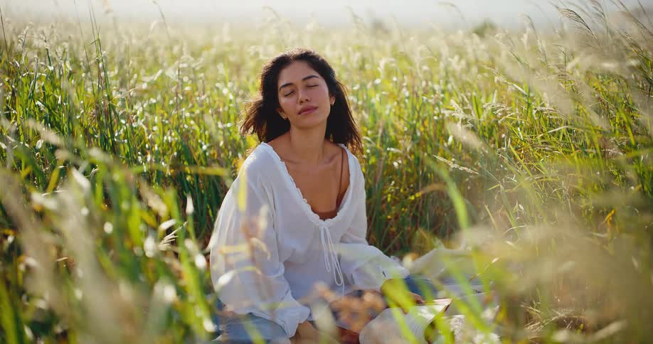A thoughtful young woman pauses from reading her book to soak in the serene atmosphere of a sunlit field of tall grass