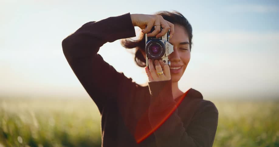 A beautiful young female photographer smiles while taking a picture with a vintage film camera in a sun-drenched grassy field