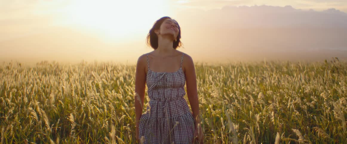 A serene young woman in a sundress enjoys a peaceful moment in a sun-drenched field of tall grass at golden hour