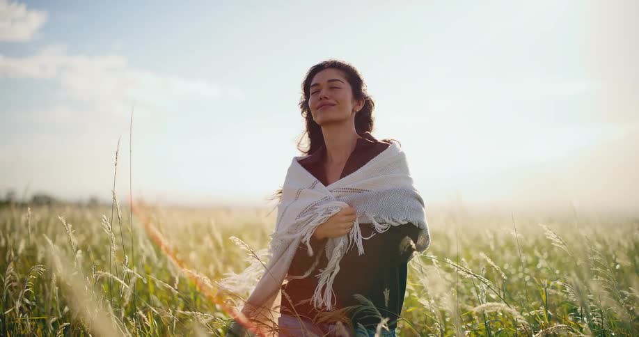 A smiling woman wraps a cozy blanket around herself while standing in a sunlit field of tall grass in slow motion