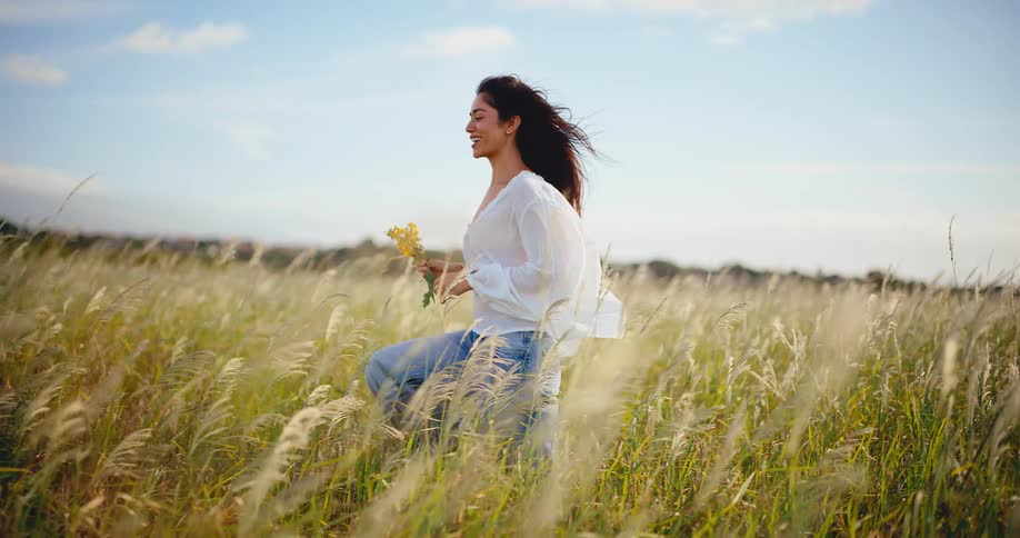 A carefree young woman holding wildflowers smiles as she walks through a windy, sunlit field of tall grass